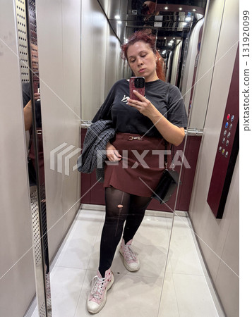 Woman taking mirror selfie in elevator wearing black t-shirt, burgundy skirt and torn tights. Urban fashion, imperfection and self-expression reflecting authenticity and casual lifestyle. 131920099