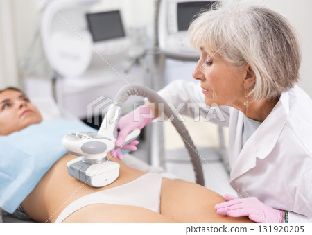 Elderly female doctor doing Rf lifting procedure on belly of young woman in beauty salon. Treatment of flabby skin 131920205