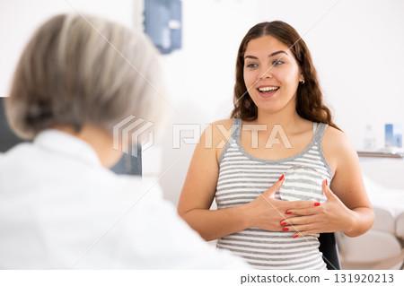 Young woman sitting in front of doctor and checking breast implant before plastic surgery in esthetic medicine center indoors 131920213