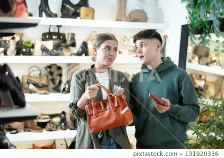 Girl contemplating handbag in store, while boyfriend shocked by price Girl contemplating handbag in store, while boyfriend shocked by price 131920336