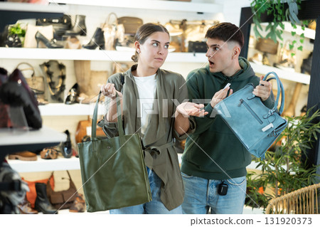 Shocked guy looking at price tag on handbag in hands of girlfriend in boutique Shocked guy looking at price tag on handbag in hands of girlfriend in boutique 131920373