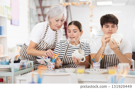 Senior woman assisting young couple painting ceramic mugs in pottery class 131920449