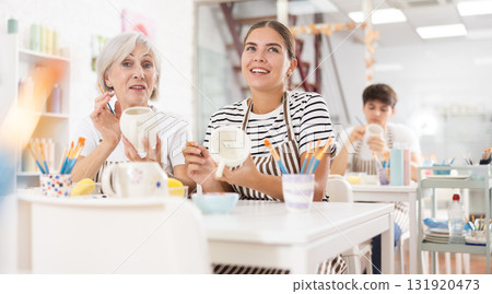 Cheerful elderly and young wowen painting mugs in pottery masterclass 131920473