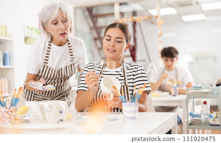 Experienced female ceramicist teaching young girl to paint ceramics in pottery studio 131920475