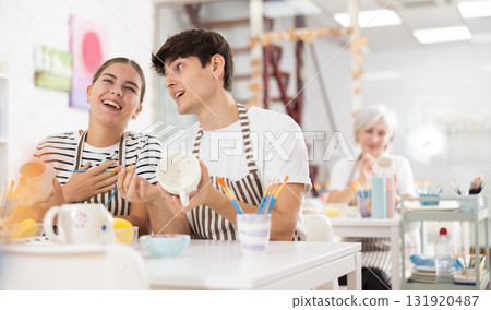Cheerful young couple decorating ceramic pottery at table in workshop 131920487