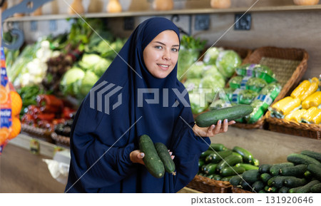 Delighted young Muslim woman purchaser choosing cucumber in grocery store Delighted young Muslim woman purchaser choosing cucumber in grocery store 131920646