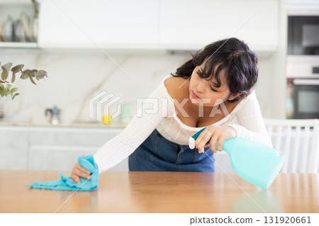 Young hardworking girl does wet cleaning in a home kitchen, wiping table with a rag Young hardworking girl does wet cleaning in a home kitchen, wiping table with a rag 131920661