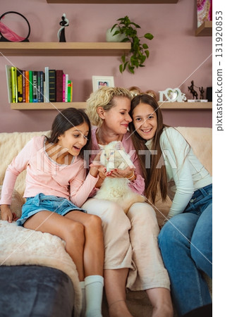 Mother and daughters cuddling cat on couch at home, enjoying family time 131920853
