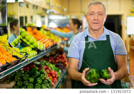 Aged salesman holding peppers at food stall in greengrocery Aged salesman holding peppers at food stall in greengrocery 131921072