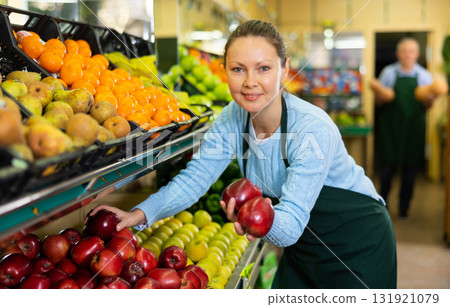 Friendly middle-aged female seller in apron arranging red apples in supermarket Friendly middle-aged female seller in apron arranging red apples in supermarket 131921079