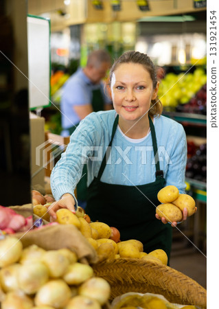Middle-aged saleswoman putting potatoes on food stall in greengrocery Middle-aged saleswoman putting potatoes on food stall in greengrocery 131921454