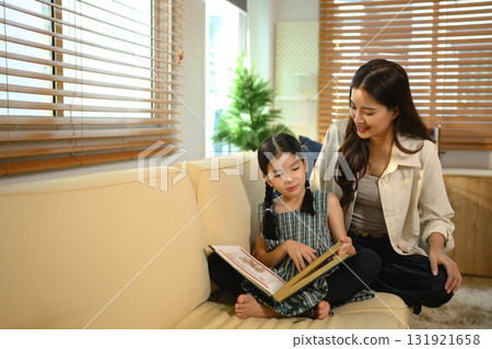 An Asian mother and daughter sit together indoors, smiling and bonding over a book 131921658