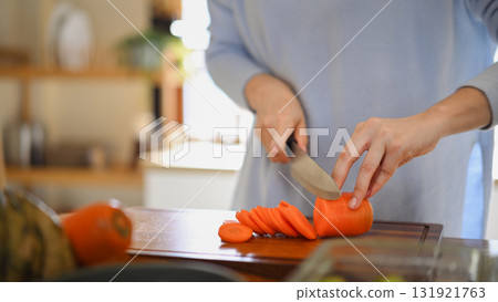Adult woman slicing fresh carrots on wooden cutting board while preparing vegetables for weekly meal prep 131921763