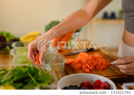 Close up of woman preparing healthy meal, placing sliced carrots into meal prep containers 131921793