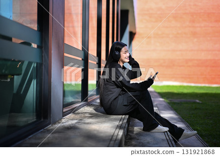 Teenage girl sitting outdoors with headphones while listening to music on her smartphone under bright sunlight 131921868
