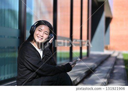 Teen girl wearing black hoodie and wireless headphones, enjoying her favorite songs outside a modern building 131921906