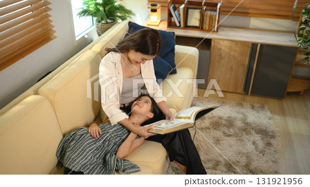 A mother reading a book to her daughter resting on her lap in the living room, enjoying a peaceful family moment 131921956