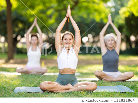 Sporty girl sitting on sports mat and making yoga meditation in lotus pose and hands up with group together in green park at daytime 131921964