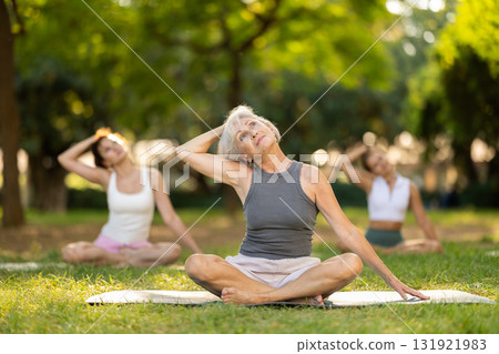 Happy old lady and class of calm sports women in activewear stretching muscles of neck while sitting in lotus pose on mats during yoga training together at city park 131921983