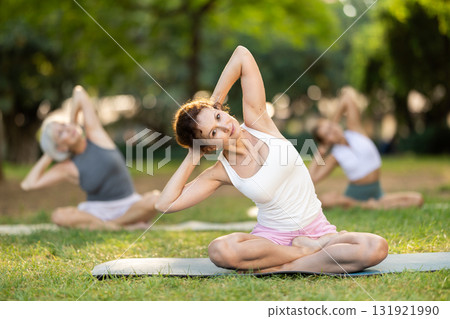 Class of calm sports women in activewear stretching muscles and doing tilts while sitting in lotus pose on mats during yoga training together at city park Class of calm sports women in activewear stretching muscles and doing tilts while sitting in lotus pose on mats during yoga training together at city park 131921990