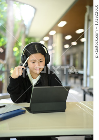 Gen Z student using tablet and stylus with headphones, smiling while studying in a modern open school environment. 131922010