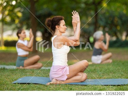 Calm european young female practicing Half Lord of fishes pose or Ardha Matsyendrasana during yoga training with group in green park 131922025