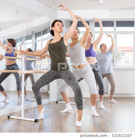 Women doing ballet at group amateurs training session in the studio performs demi plie near the ballet barre, standing in ballet stance 131922107