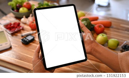 Person holding digital tablet with blank screen on kitchen counter surrounded by fresh fruits and vegetables for healthy lifestyle concept Person holding digital tablet with blank screen on kitchen counter surrounded by fresh fruits and vegetables for healthy lifestyle concept 131922153
