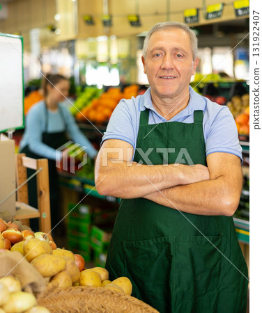 Portrait of confident mature male supermarket worker wearing apron standing in middle of grocery store 131922407