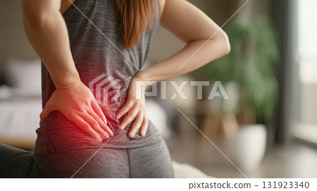 Woman experiencing lower back pain while sitting on a yoga mat in a modern living room setting 131923340