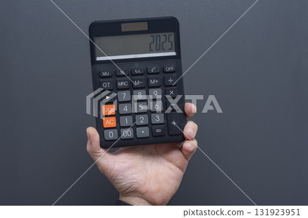 Man hand holding black digital calculator displaying 2025 on LCD screen against gray background. Business finance and annual planning concept for new year calculations and budgeting. 131923951
