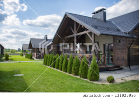 Blurred modern wooden house exterior, nestled amidst lush green grass and manicured bushes under a bright sky. 131923956