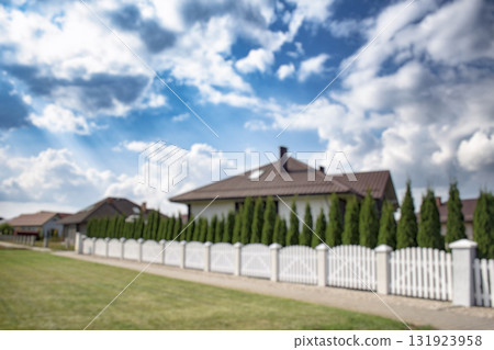 Blurred view of a quiet suburban neighborhood with private houses, white fences, and a curved street under a cloudy sky. Background for real estate, property, and urban development themes. 131923958