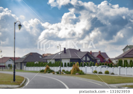 Blurred view of a quiet suburban neighborhood with private houses, white fences, and a curved street under a cloudy sky. Background for real estate, property, and urban development themes. 131923959