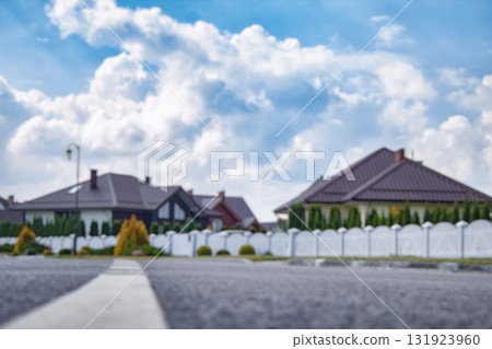 Blurred view of a quiet suburban neighborhood with private houses, white fences, and a curved street under a cloudy sky. Background for real estate, property, and urban development themes. Blurred view of a quiet suburban neighborhood with private houses, white fences, and a curved street under a cloudy sky. Background for real estate, property, and urban development themes. 131923960
