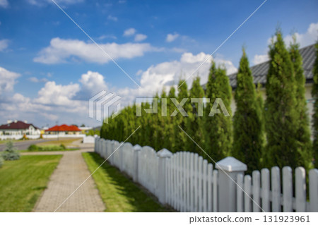 Blurred view of a quiet suburban neighborhood with private houses, white fences, and a curved street under a cloudy sky. Background for real estate, property, and urban development themes. 131923961
