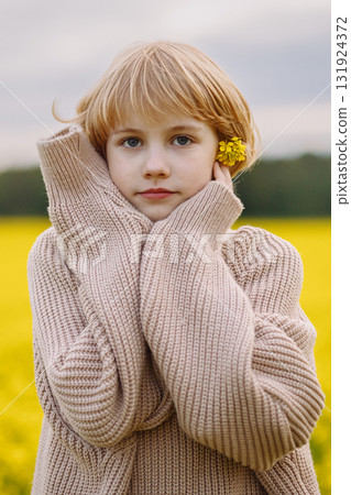 Blonde girl holding yellow flower near face in beige sweater, front view 131924372