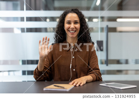 Woman smiling and waving during a friendly virtual meeting, wearing earphones at her desk, connecting with remote colleagues in a casual, professional video call setting Woman smiling and waving during a friendly virtual meeting, wearing earphones at her desk, connecting with remote colleagues in a casual, professional video call setting 131924955