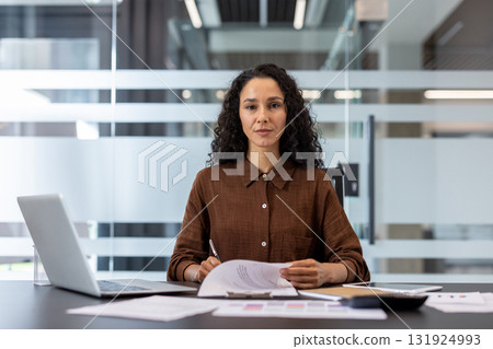 Professional businesswoman working at her desk with a laptop and documents, holding a pen and looking directly at the viewer with a confident and serious expression 131924993