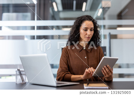 Professional businesswoman working at her desk, concentrating while using a digital tablet for tasks and checking information, with a laptop nearby in a contemporary office setting 131925003