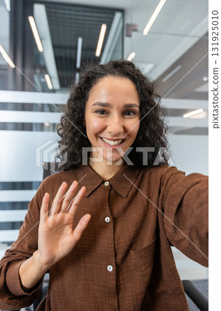 Young woman with curly hair smiling and waving during a virtual meeting or online conference, communicating remotely from a modern business office environment 131925010