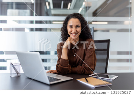 Confident young businesswoman smiling and looking at the camera, posing at a modern office desk with a laptop, reflecting professionalism and success in a corporate setting 131925027
