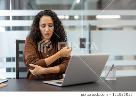 Businesswoman feeling discomfort in her elbow while working on a laptop at a desk in a modern office, experiencing joint pain from prolonged computer use 131925035