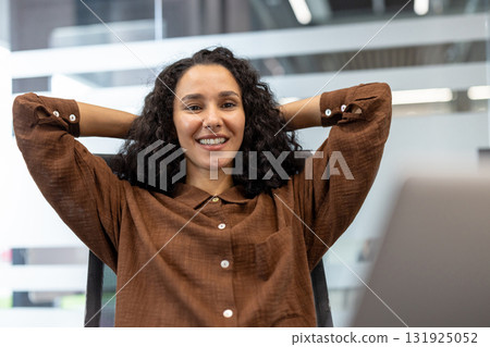 Confident young business woman with curly hair relaxing at her desk in a modern office, smiling genuinely and feeling comfortable after completing her work tasks Confident young business woman with curly hair relaxing at her desk in a modern office, smiling genuinely and feeling comfortable after completing her work tasks 131925052