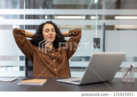 Young woman with curly hair leaning back at her desk, eyes closed and hands behind head, enjoying a calm, stress-free break during a busy remote workday at home Young woman with curly hair leaning back at her desk, eyes closed and hands behind head, enjoying a calm, stress-free break during a busy remote workday at home 131925055