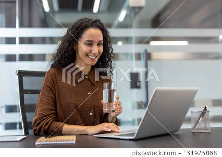 Smiling woman engaged in a virtual conference or online meeting, holding a glass of water, while working on a laptop at her contemporary office desk, representing modern communication and wellness 131925068