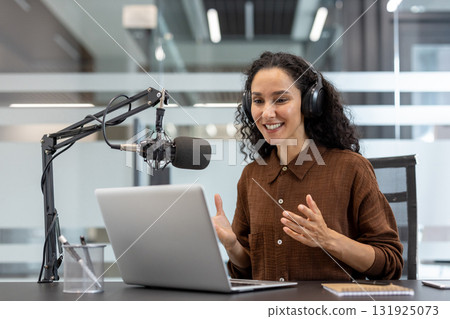 Woman wearing headphones and actively engaging in an online podcast or webinar, speaking into a professional microphone and gesturing while looking at her laptop screen in a modern office Woman wearing headphones and actively engaging in an online podcast or webinar, speaking into a professional microphone and gesturing while looking at her laptop screen in a modern office 131925073