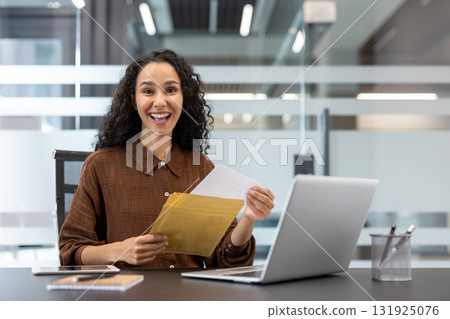 Young professional woman at her office desk opens an envelope with a surprised, joyful smile, celebrating promotion news or a positive financial outcome while working on her laptop Young professional woman at her office desk opens an envelope with a surprised, joyful smile, celebrating promotion news or a positive financial outcome while working on her laptop 131925076