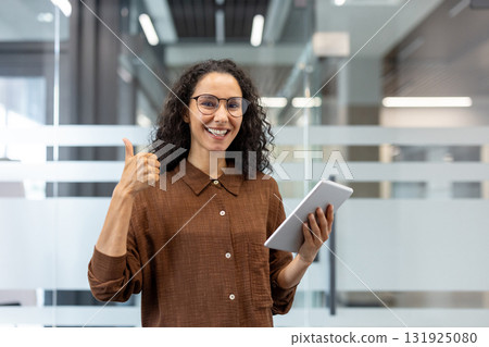 Happy businesswoman giving thumbs up gesture while holding a digital tablet and smiling, standing in a modern office, showing approval, success, and confidence in her work Happy businesswoman giving thumbs up gesture while holding a digital tablet and smiling, standing in a modern office, showing approval, success, and confidence in her work 131925080