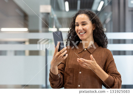 Woman standing in a modern office using a smartphone for a video call, smiling and gesturing while communicating remotely, representing business connection and corporate technology 131925083